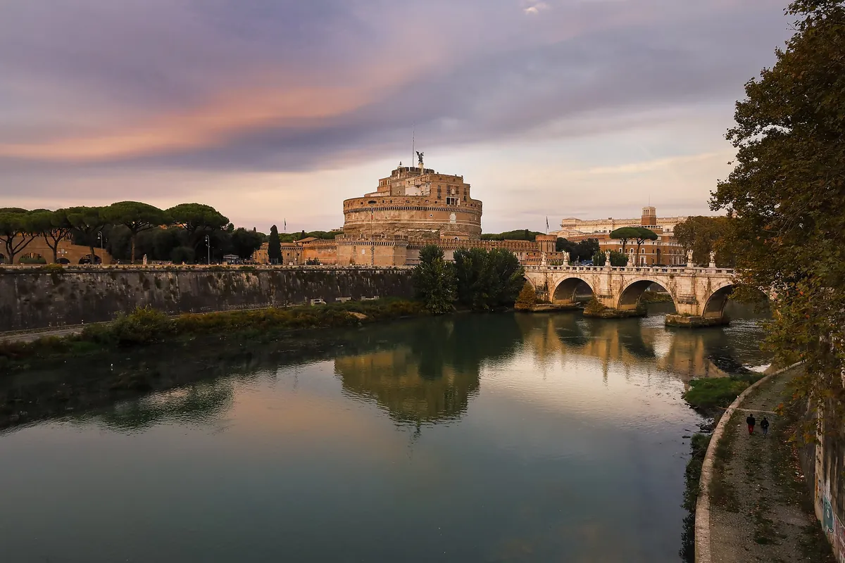 Castel Sant'Angelo and its bridge over the Tiber at sunset, with pink clouds above and the round fortress reflected in the river; Act 3 of Tosca takes place on its upper levels.